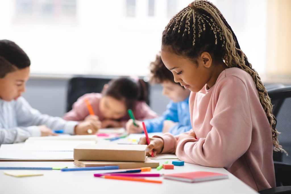 Portrait of small black girl sitting at desk in classroom Small girl with braids sitting at table in classroom, writing or drawing in notebook