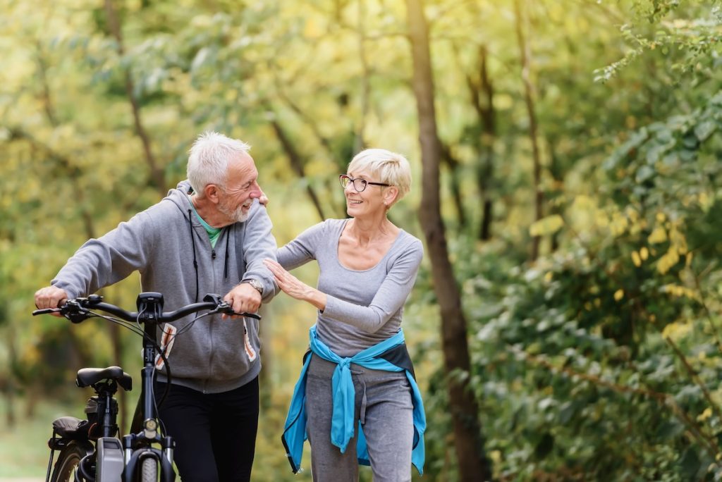 Cheerful active senior couple with bicycles walking through park together. Perfect activities for elderly people. Cheerful active senior couple with bicycles walking through park together.