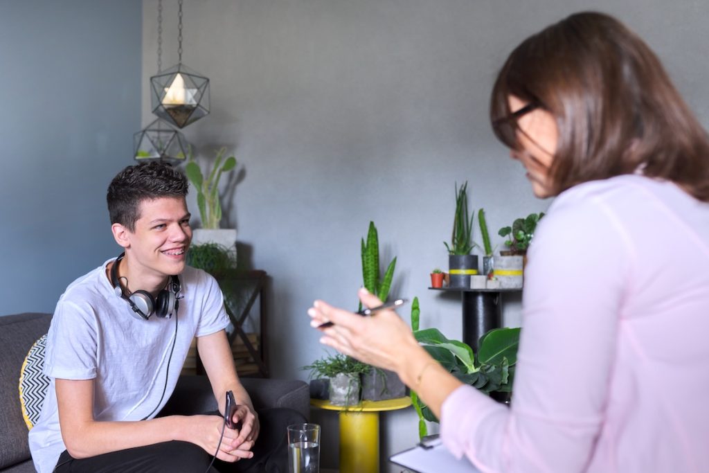 A smiling teenage boy with headphones around his neck sits on a couch, engaged in conversation with a therapist in a room decorated with plants.