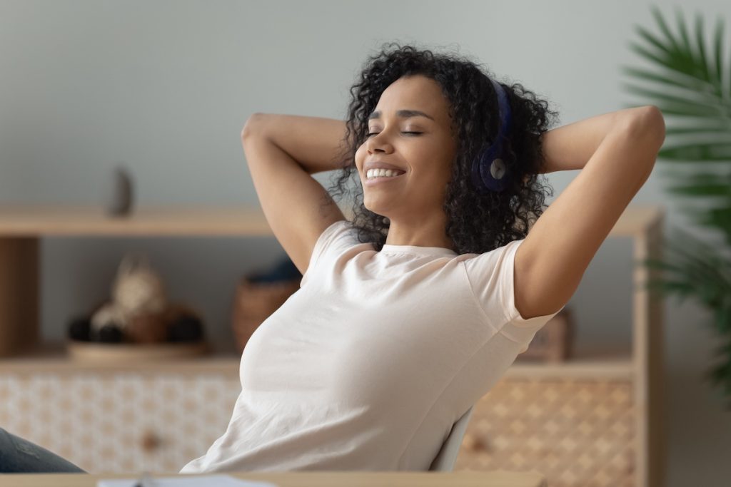 Serene african woman closed eyes enjoy music through wireless headphones Serene african woman put hands behind head closed eyes enjoy music through wireless headphones smiling enjoy favourite music at home or workplace, mood and hobby, stress relief and relaxation concept