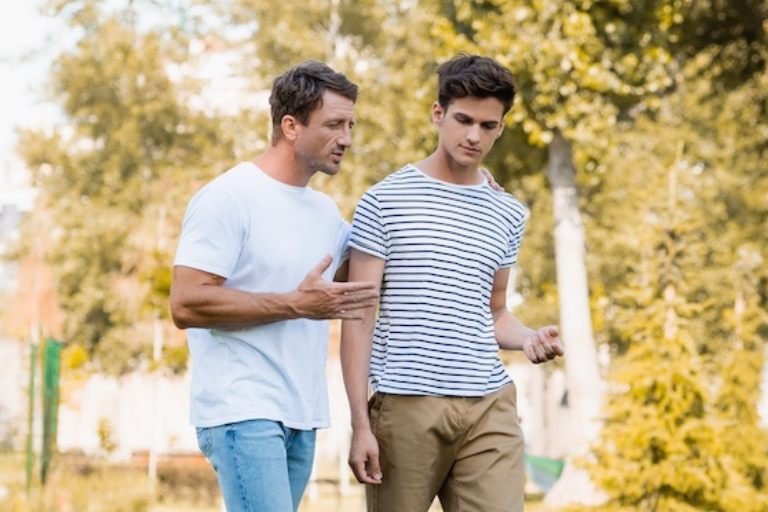 panoramic shot of father walking and talking with teenager son in park