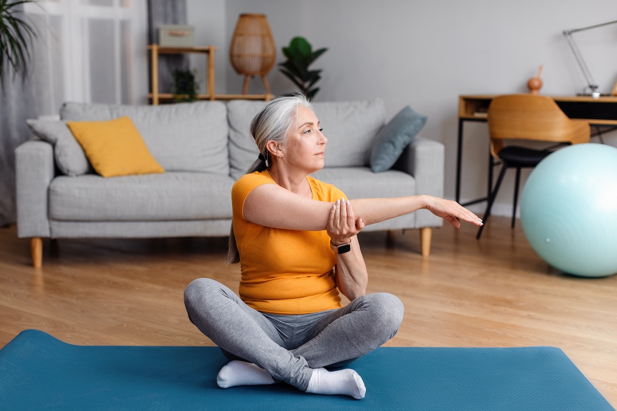 Active senior woman stretching her arm on mat at home, doing flexibility exercises, practicing yoga or pilates indoors after stroke.