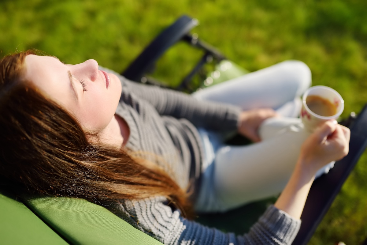 Woman relaxing with closed eyes and cup of coffee in lounge chair on a sunny day outdoors, daydreaming.