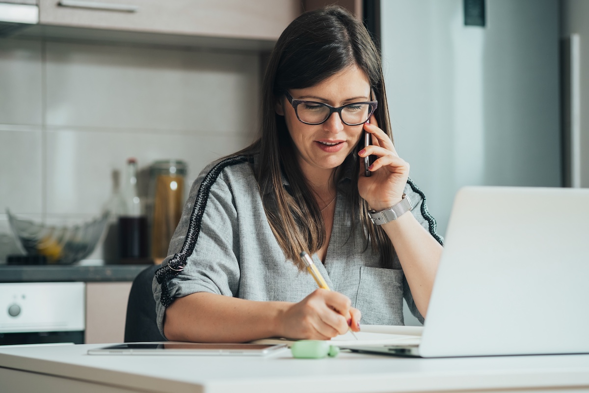 Woman on phone call taking notes at home desk with laptop