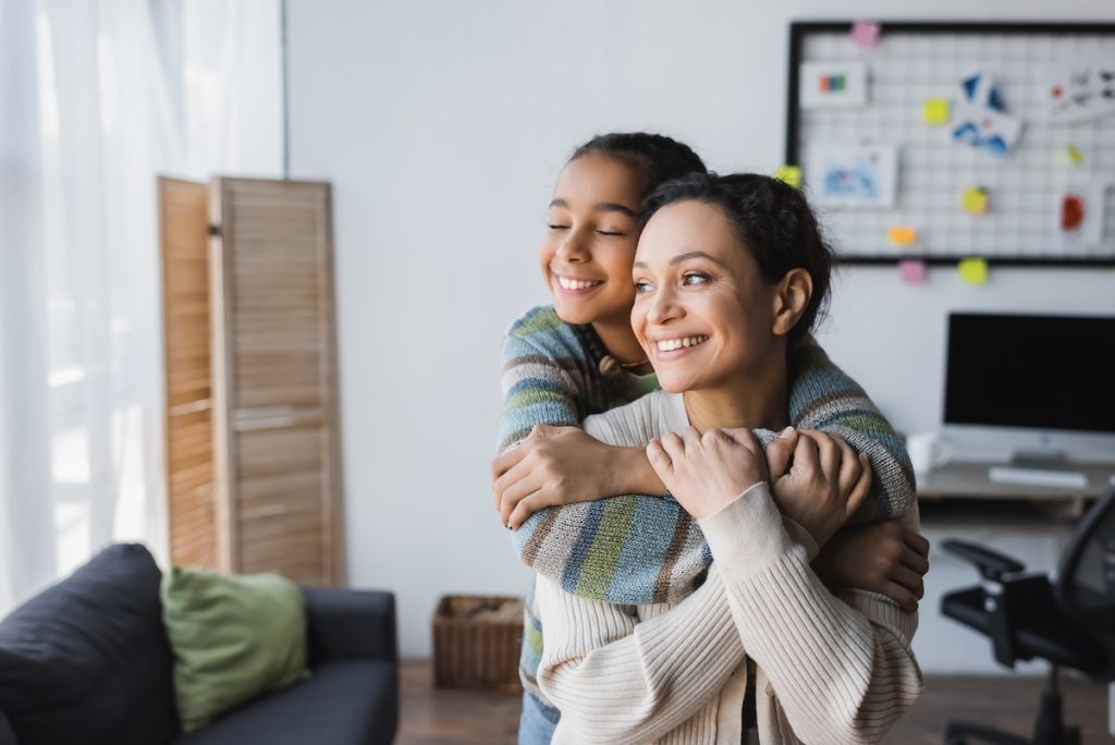 smiling african american girl with closed eyes hugging happy mom near computer monitor on blurred background